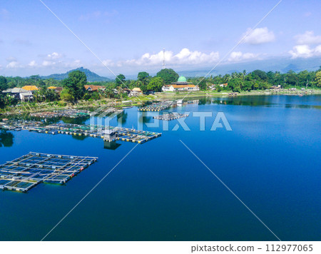 An aerial view of a small lakeside town with a mosque, surrounded by trees and houses. Fish cages are visible in the water. An aerial view of a small lakeside town with a mosque, surrounded by trees and houses. Fish cages are visible in the water. 112977065