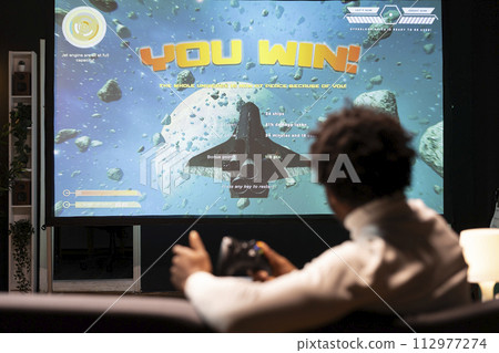 Man looking at winning screen of science fiction game on large smart TV. African american gamer receiving victory message after completing all videogame levels, watching statistics roll on display 112977274