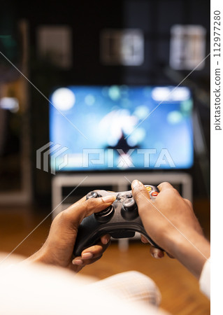 Focus shot on man holding controller in apartment, playing videogames on smart TV display in blurry background. Gamer on sofa using joypad to participate in game on console attached to television set 112977280