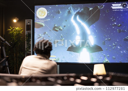 BIPOC man plays science fiction singleplayer game with overlay HUD showing health bars and minimap on huge TV screen. Player enjoying high quality ray tracing graphics in console videogame BIPOC man plays science fiction singleplayer game with overlay HUD showing health bars and minimap on huge TV screen. Player enjoying high quality ray tracing graphics in console videogame 112977281