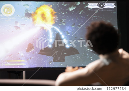 Gamer playing classic arcade action videogame on ultrawide smart TV, flying through space debris. Man enjoying leisure time at home using high tech gaming console and controller to solve missions Gamer playing classic arcade action videogame on ultrawide smart TV, flying through space debris. Man enjoying leisure time at home using high tech gaming console and controller to solve missions 112977284