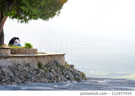 man and a woman sit near a tree against a background of travertines in Pamukkale 112977455