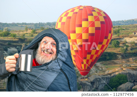Happy bearded man in a sleeping bag with a cup of coffee against the backdrop of nature 112977456
