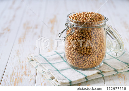Raw soybeans in glass storage jar on a white wooden table. Raw soybeans in glass storage jar on a white wooden table. 112977536