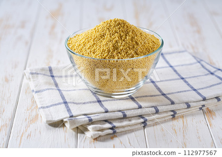 Raw grains of millet in a glass plate on a white wooden table. Raw grains of millet in a glass plate on a white wooden table. 112977567
