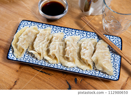 Portion of gyoza dumplings served with bowl of soy sauce and chopsticks. Traditional oriental culinary art, acquaintance with culture through national cuisine Portion of gyoza dumplings served with bowl of soy sauce and chopsticks. Traditional oriental culinary art, acquaintance with culture through national cuisine 112980650