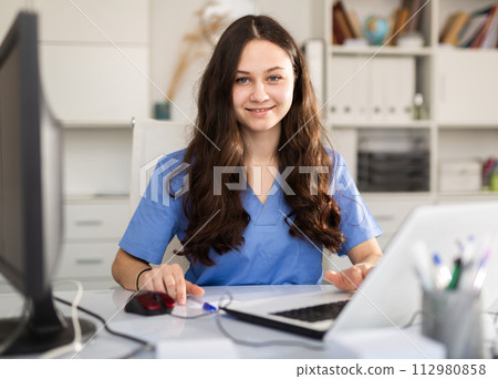 Young female trainee doctor is sitting at workplace at a computer Young female trainee doctor is sitting at workplace at a computer 112980858