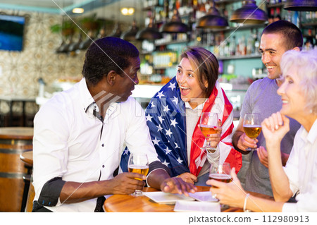 Cheerful multiracial footbal fans waving the flag of USA while drinking beer and watching tournament in sport bar Cheerful multiracial footbal fans waving the flag of USA while drinking beer and watching tournament in sport bar 112980913