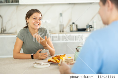 Cheerful young girl talking with boyfriend over cup of coffee in home kitchen Cheerful young girl talking with boyfriend over cup of coffee in home kitchen 112981155