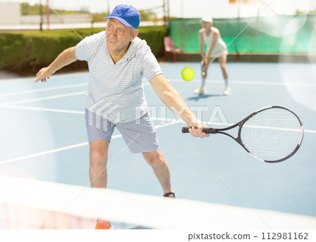 Man with black racket playing match in open behind net court outdoors. 112981162