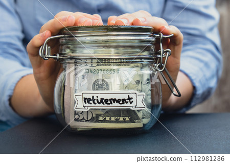 Unrecognizable woman holding Glass jar full of American currency dollars cash banknote with text RETIREMENT. Preparation saving money. Moderate consumption and economy Unrecognizable woman holding Glass jar full of American currency dollars cash banknote with text RETIREMENT. Preparation saving money. Moderate consumption and economy 112981286