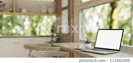 A laptop computer mockup on a wooden dining table against the window in a neutral modern kitchen. A laptop computer mockup on a wooden dining table against the window in a neutral modern kitchen. 112982008