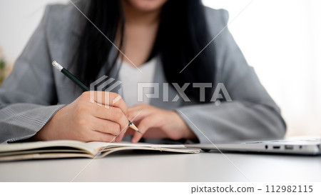 A cropped shot of a businesswoman focused on writing or taking notes in the notebook with a pencil. A cropped shot of a businesswoman focused on writing or taking notes in the notebook with a pencil. 112982115