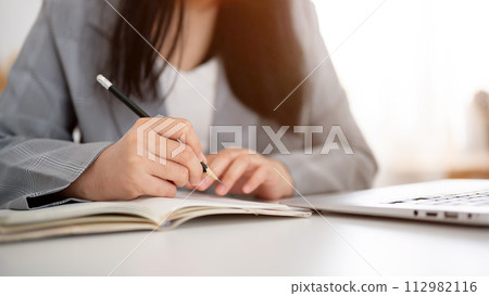 A cropped shot of a businesswoman focused on writing or taking notes in the notebook with a pencil. A cropped shot of a businesswoman focused on writing or taking notes in the notebook with a pencil. 112982116