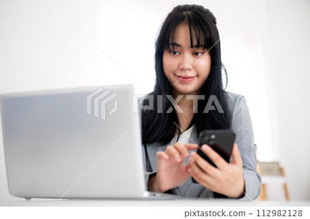 A young Asian businesswoman is looking at her laptop screen while using her smartphone at her desk. 112982128