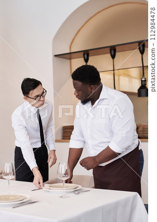 Vertical medium long shot of Asian headwaiter and African American busser discussing work while setting table in modern restaurant 112984478