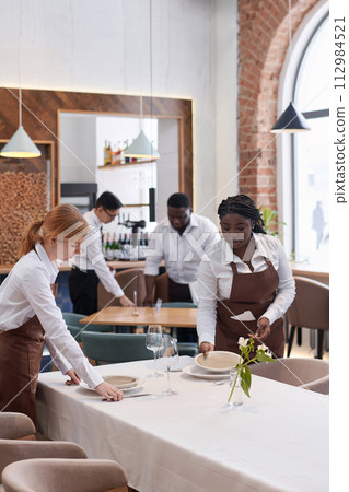 Vertical shot of ethnically diverse waiting team working in modern restaurant setting tables in morning Vertical shot of ethnically diverse waiting team working in modern restaurant setting tables in morning 112984521