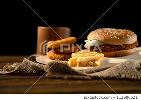 Gourmet burger with onion rings and side of fries staged on wood with a rustic feel and inviting lighting. Gourmet burger with onion rings and side of fries staged on wood with a rustic feel and inviting lighting. 112986621