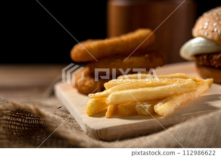 Selective focus on golden French fries with onion rings and a burger in the backdrop, on a wooden board. Selective focus on golden French fries with onion rings and a burger in the backdrop, on a wooden board. 112986622