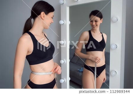 Woman in underwear measures her body with tape measure in front of the mirror. 112989344