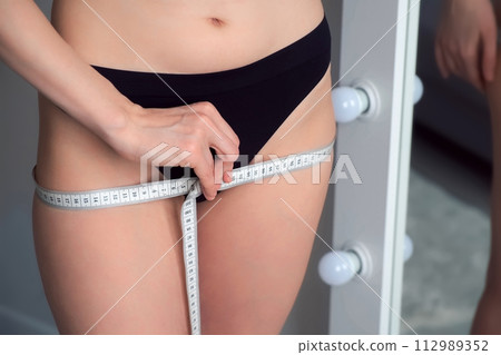 Woman in underwear measures her body with tape measure in front of the mirror. 112989352