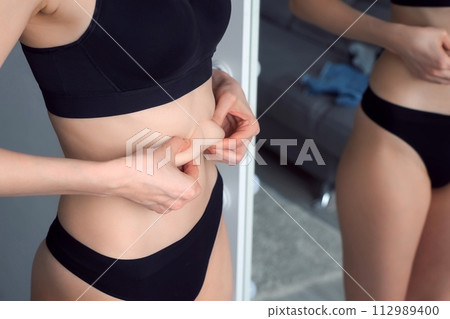 A young thin woman in black underwear critically examines her figure in mirror. A young thin woman in black underwear critically examines her figure in mirror. 112989400