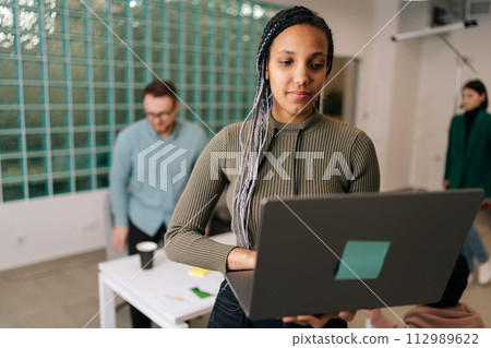 Portrait of African female entrepreneur standing in modern office holding laptop computer, looking to screen. Diverse businesspeople negotiating on blurred background on workplace 112989622