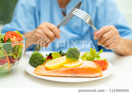 Asian elderly woman patient eating Salmon steak breakfast with vegetable healthy food while sitting and hungry on bed in hospital. 112990080