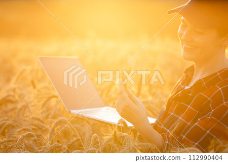 Female farmer working with laptop on wheat field. Smart farming and digital agriculture.. Female farmer working with laptop on wheat field. Smart farming and digital agriculture.. 112990404