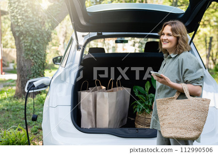 Woman with shopping bag next to a charging electric car in the yard of a country house Woman with shopping bag next to a charging electric car in the yard of a country house 112990506