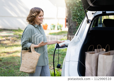 Woman with shopping bag next to a charging electric car in the yard of a country house Woman with shopping bag next to a charging electric car in the yard of a country house 112990510