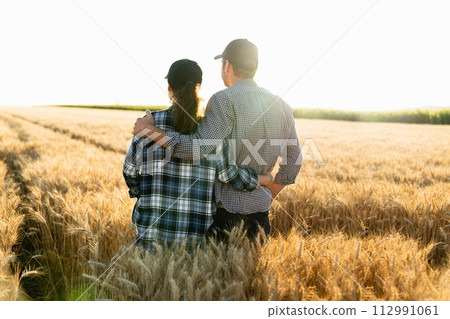 A couple of farmers in plaid shirts and caps stand embracing on agricultural field of wheat at sunset. A couple of farmers in plaid shirts and caps stand embracing on agricultural field of wheat at sunset. 112991061