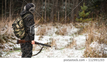 Male hunter in camouflage and with backpack, armed with a rifle, walks through the snowy winter forest Male hunter in camouflage and with backpack, armed with a rifle, walks through the snowy winter forest 112991283