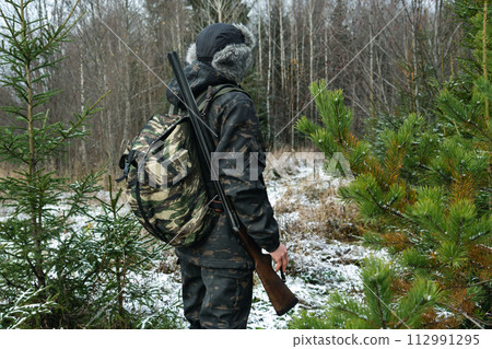 Male hunter in camouflage and with backpack, armed with a rifle, walks through the snowy winter forest Male hunter in camouflage and with backpack, armed with a rifle, walks through the snowy winter forest 112991295