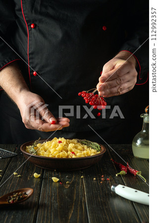Adding viburnum berries in a cook hand to a plate of pasta. The chef at the kitchen table in the tavern is preparing a delicious lunch 112992357
