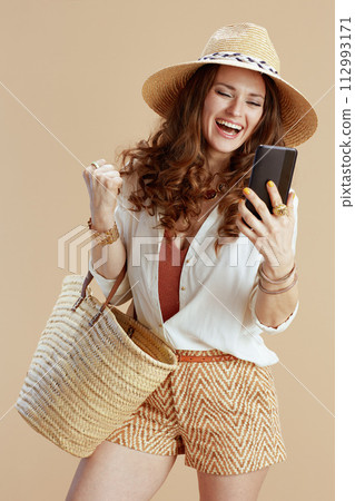 smiling woman using phone with raised arms rejoicing on beige 112993171