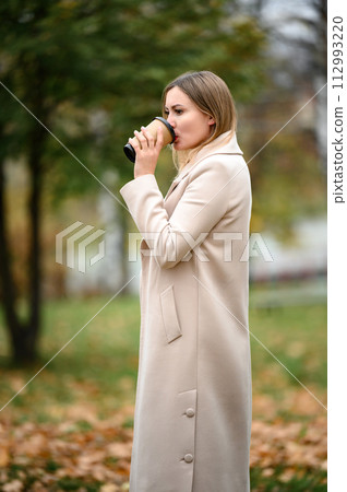 A young girl in a long autumn coat drinks coffee in an autumn park. A young girl in a long autumn coat drinks coffee in an autumn park. 112993220