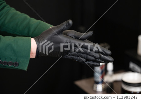 A barber in black gloves rubs wax and pomade for styling a clients hair. 112993242