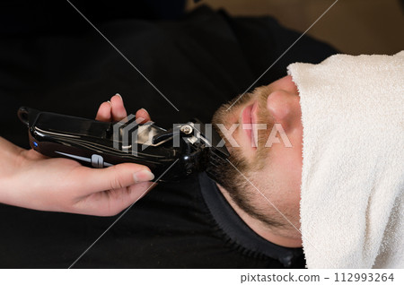 The clients head is covered with a towel during a beard trim. Shortening the length of the beard from the sides by the master for the client. 112993264