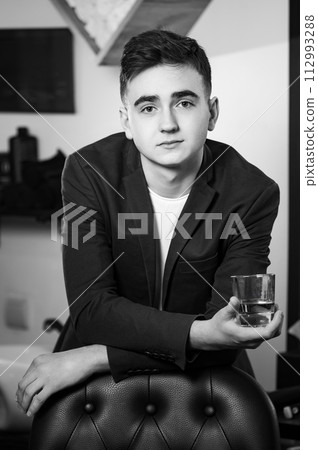 Portrait of a young barber in a jacket and white t-shirt. The client leaned on the barber's chair, holding a glass of whiskey in his hands. Portrait of a young barber in a jacket and white t-shirt. The client leaned on the barber's chair, holding a glass of whiskey in his hands. 112993288