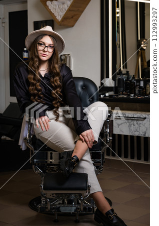 Portrait of a young hairdresser girl in a hat. A female barber is sitting in a chair and waiting for a client. Portrait of a young hairdresser girl in a hat. A female barber is sitting in a chair and waiting for a client. 112993297