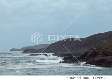 Moody view of Scourie Bay with tumultuous waves crashing against rugged cliffs under somber sky Moody view of Scourie Bay with tumultuous waves crashing against rugged cliffs under somber sky 112994099