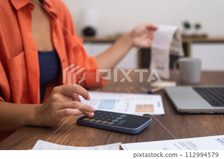 Expense Management: Seated at a desk with a laptop, a woman uses her smartphone calculator to compute household finances Expense Management: Seated at a desk with a laptop, a woman uses her smartphone calculator to compute household finances 112994579