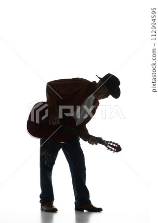 Silhouette of man in fedora hat playing guitar, performing isolated on white background. Black and white image. 112994595