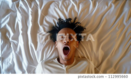 Man lying on white bed, open wide mouth in room alone at apartment 112995660
