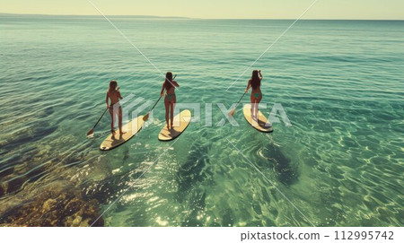 Three happy women stands on a sup boards. In the background, the ocean and the sunset 112995742