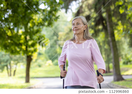 Satisfied and active senior gray-haired woman is doing sports and Nordic walking in the park, holding trekking poles in her hands, looking to the side. 112996860
