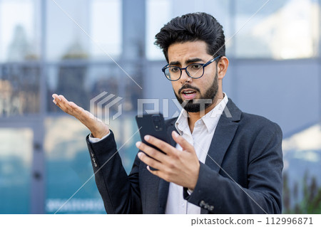 Close-up photo of a shocked young Muslim businessman standing outside on the street and looking at the phone screen in surprise. 112996871