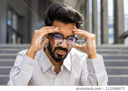 A close-up photo of a young Indian male office worker sitting outside a building on the steps and holding his head in his hands, feeling severe pain and pressure. 112996873