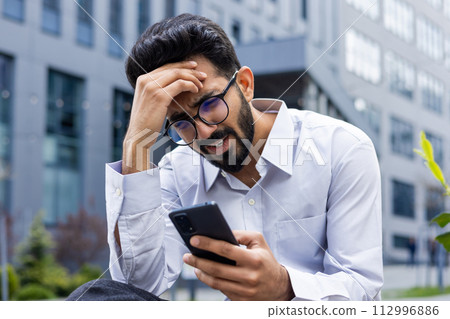 Close-up photo of shocked and upset young Indian man sitting outside holding head with hand and looking at phone screen after receiving bad news. Close-up photo of shocked and upset young Indian man sitting outside holding head with hand and looking at phone screen after receiving bad news. 112996886